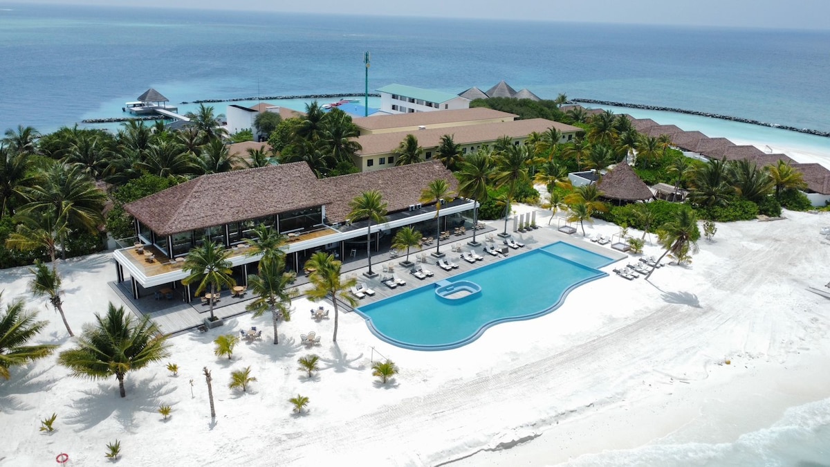 An aerial view reveals a resort with a large pool surrounded by lounge chairs and palm trees. The sandy beach leads to clear blue waters. Nearby buildings and thatched-roof structures are visible against the vibrant greenery, contributing to a tranquil coastal setting.