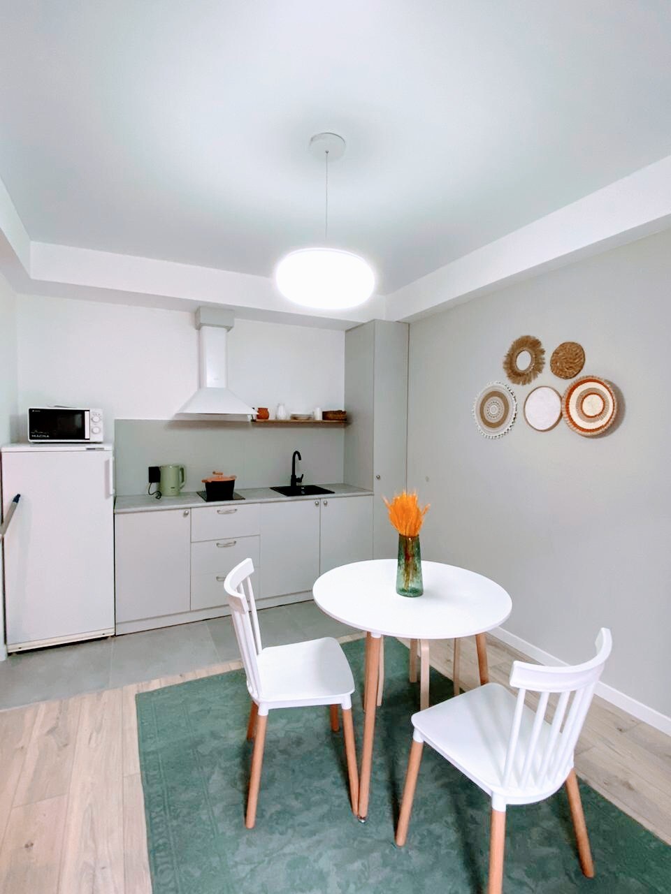 A modern kitchen area is presented, featuring white cabinetry and a contrasting dark sink. A round table with three white chairs is positioned on a soft green rug. Decorative plates adorn the wall, complemented by a vase with decorative branches at the center of the table.