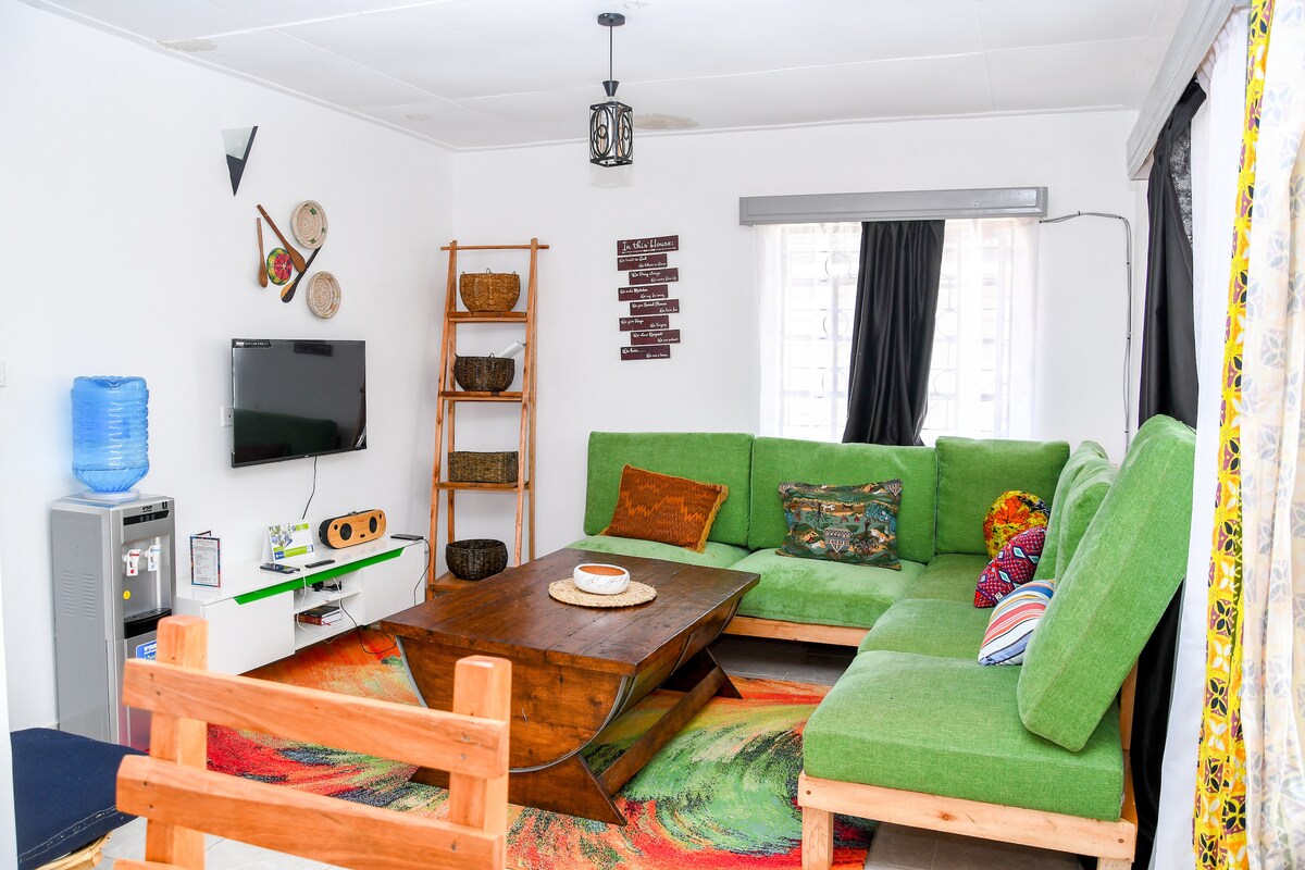 A bright and inviting living area features a large green sectional sofa adorned with colorful cushions. A wooden coffee table sits centrally on a patterned rug. Decorative wall art and woven baskets add character, while a TV is mounted on a white wall opposite the seating.