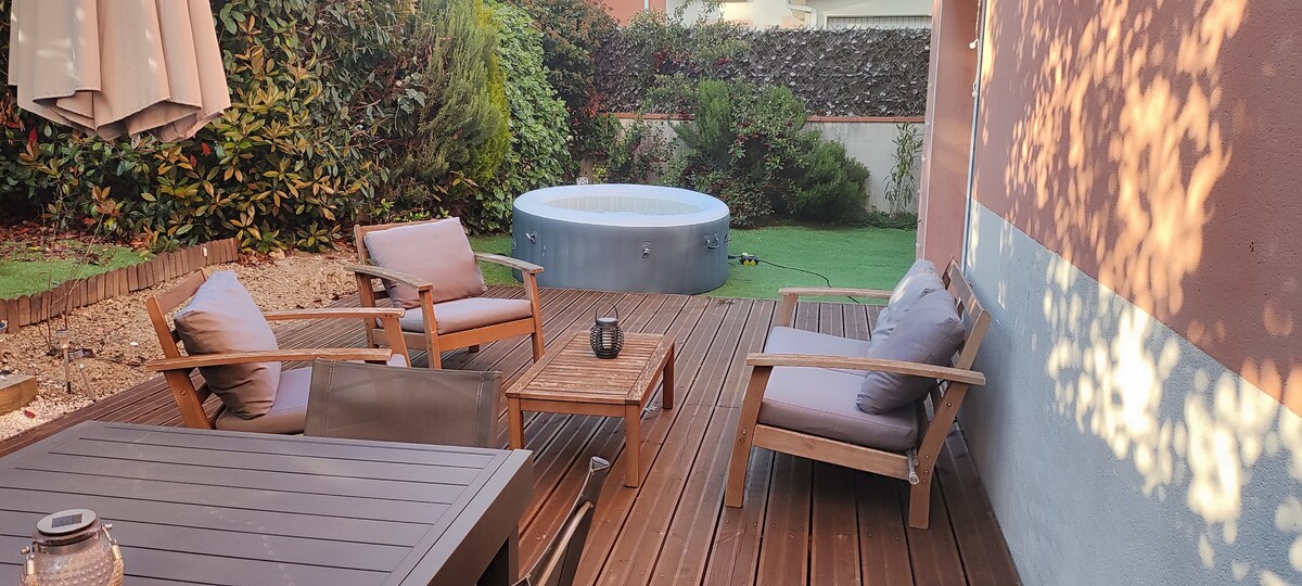 An outdoor terrace features wooden decking, equipped with a table and four wooden chairs. Two lounge chairs provide comfortable seating. A spa is positioned nearby, surrounded by greenery. Soft shadows play across the wall and ground, creating a tranquil setting.