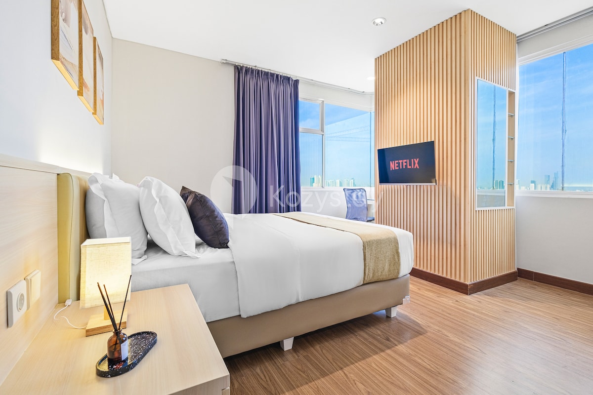The master bedroom features a queen-size bed with a neutral-toned bedspread and decorative pillows. A 32" Smart TV is mounted on the wall, with access to Netflix. Natural light fills the room through large windows, dressed with dark curtains.