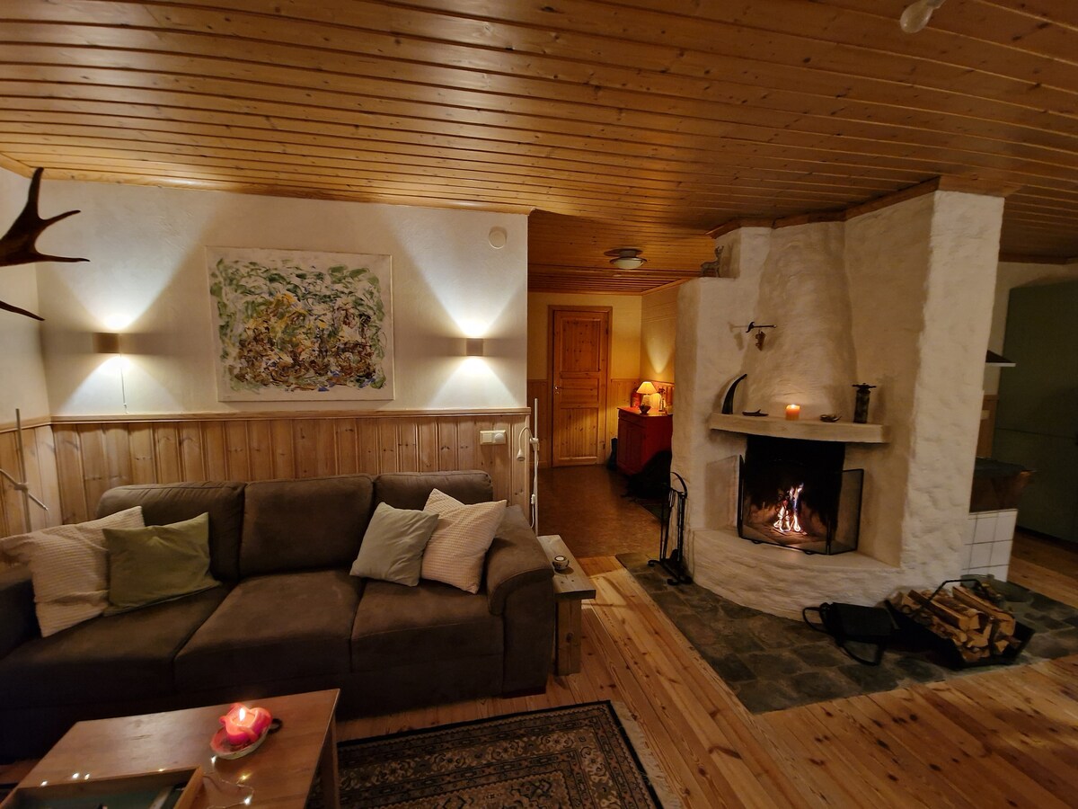 A cozy living area is shown, featuring a spacious sofa adorned with soft pillows and a wooden coffee table. A stone fireplace stands prominently, providing a warm glow. The light from wall sconces complements the natural wood ceiling and warm tones of the surrounding decor.