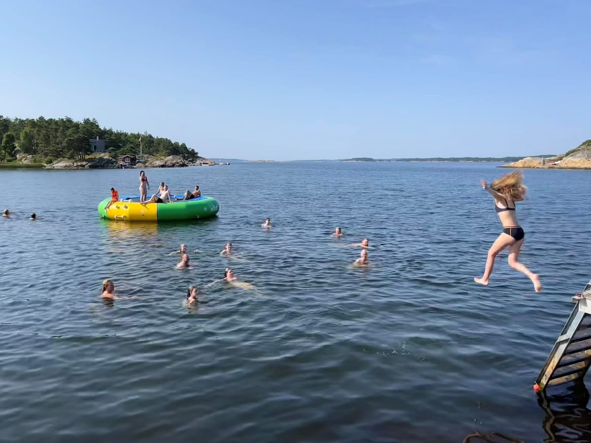 A sunny outdoor scene captures individuals swimming in calm waters, while others enjoy a colorful floating trampoline. A person is seen mid-air, preparing to jump into the water. Lush greenery and rocky formations frame the background, contributing to the serene coastal setting.