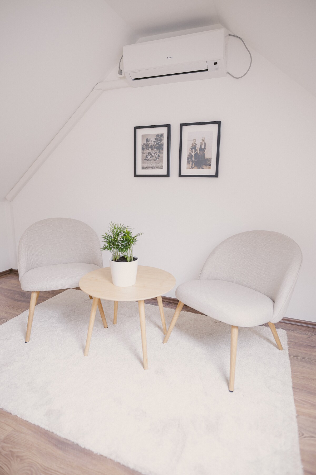 A cozy seating area features two upholstered chairs with light gray fabric, arranged around a round wooden table. A small potted plant adds a touch of greenery, while two framed artworks hang on the white wall above. A soft rug enhances the welcoming atmosphere.