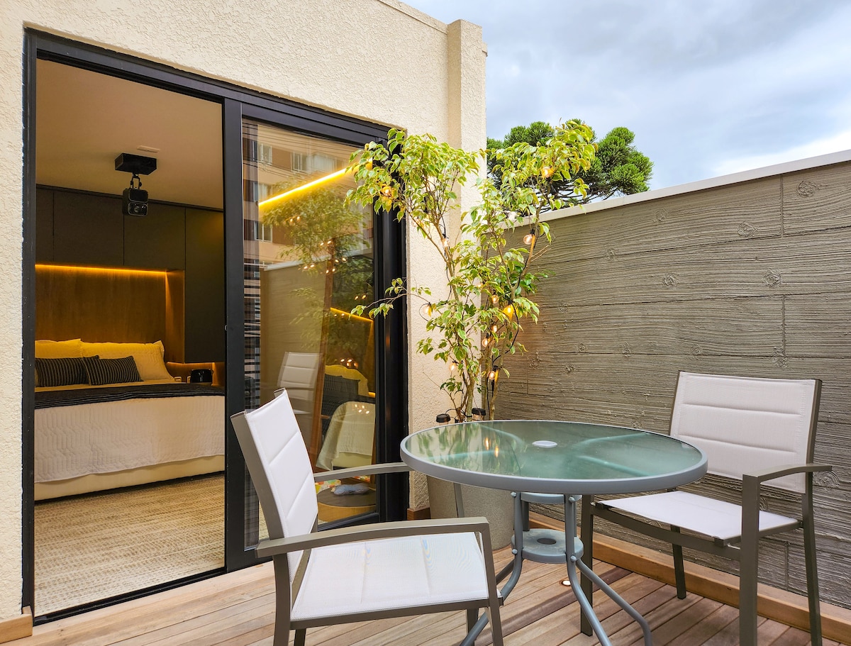 An outdoor space features a round glass table surrounded by four white chairs, creating a cozy dining area. A small plant adds greenery to the setting. The backdrop shows sliding glass doors leading to the interior, where a well-appointed bedroom can be seen.