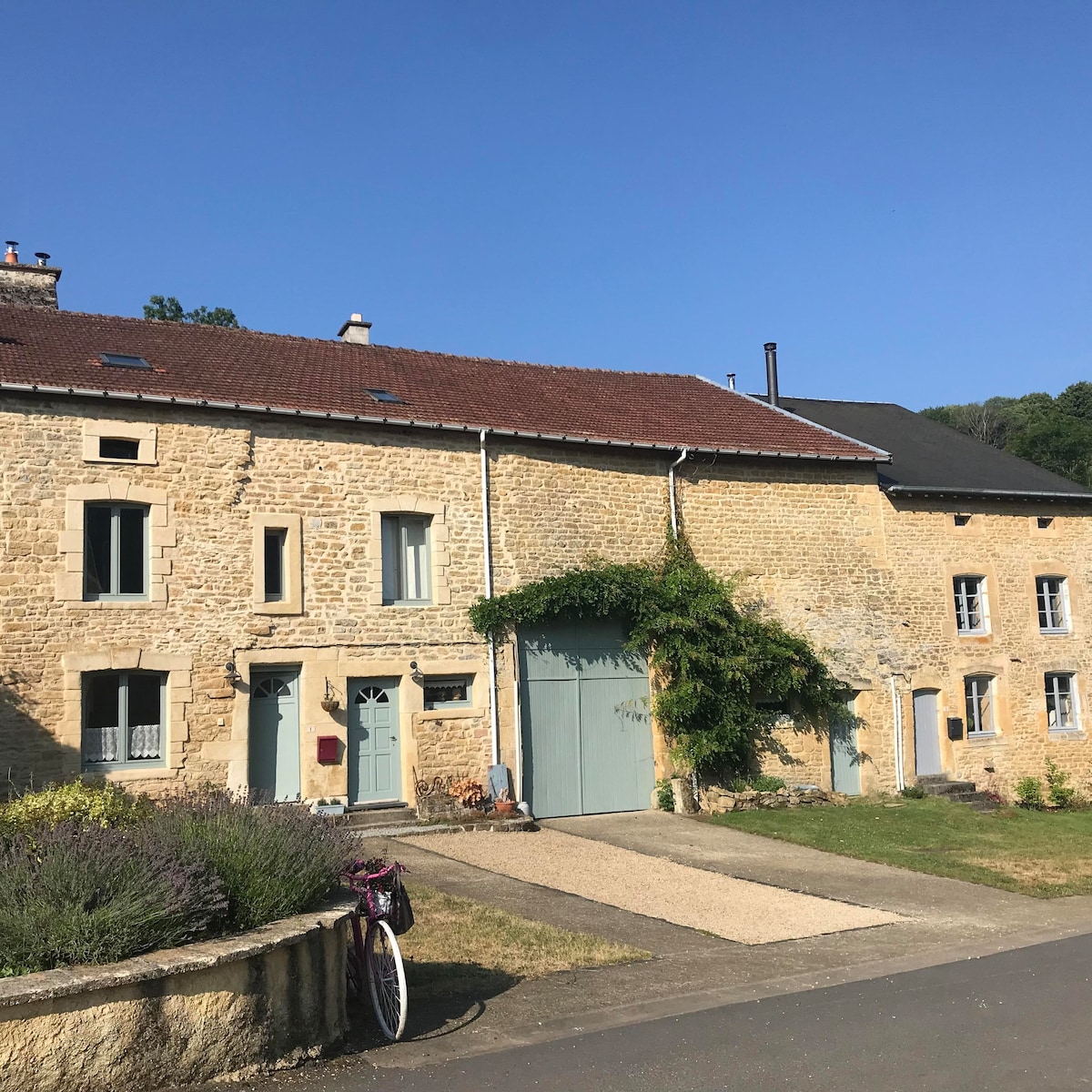 A charming stone building stands against a clear blue sky. The structure features a mix of light and darker stone, with large windows and a green door. Lush greenery accompanies the entrance, while a bicycle rests near the gravel driveway.