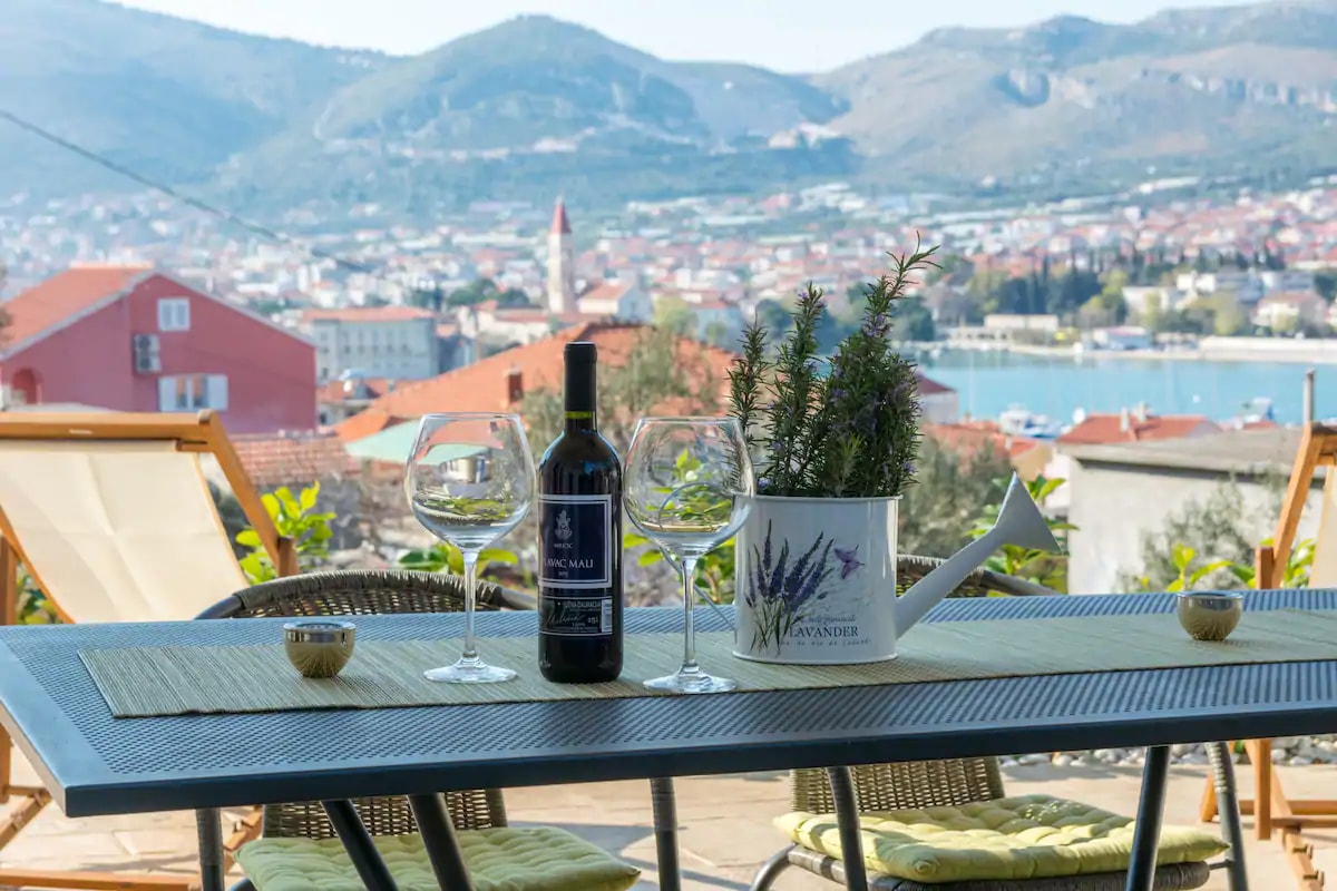 An outdoor terrace is highlighted by a dining table set with glasses and a bottle of wine. A decorative planter adds greenery, while the picturesque backdrop features the historic town of Trogir and the Adriatic Sea, visible through a clear view of the landscape.