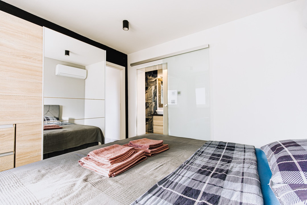 A modern bedroom features a spacious bed adorned with neatly folded towels. Mirrored cabinetry reflects the room's sleek design. A glass sliding door leads to the bathroom area, and natural light fills the space from a nearby window.