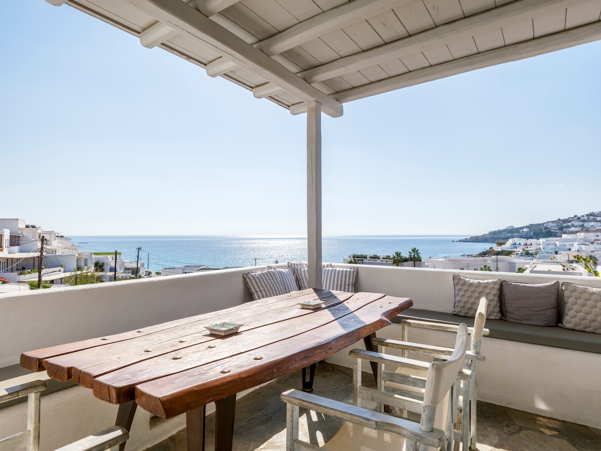 A spacious outdoor terrace features a large wooden dining table surrounded by white chairs. Soft cushions add comfort to the seating area. The tranquil Aegean Sea is visible in the background, with sunlight shimmering on its surface.