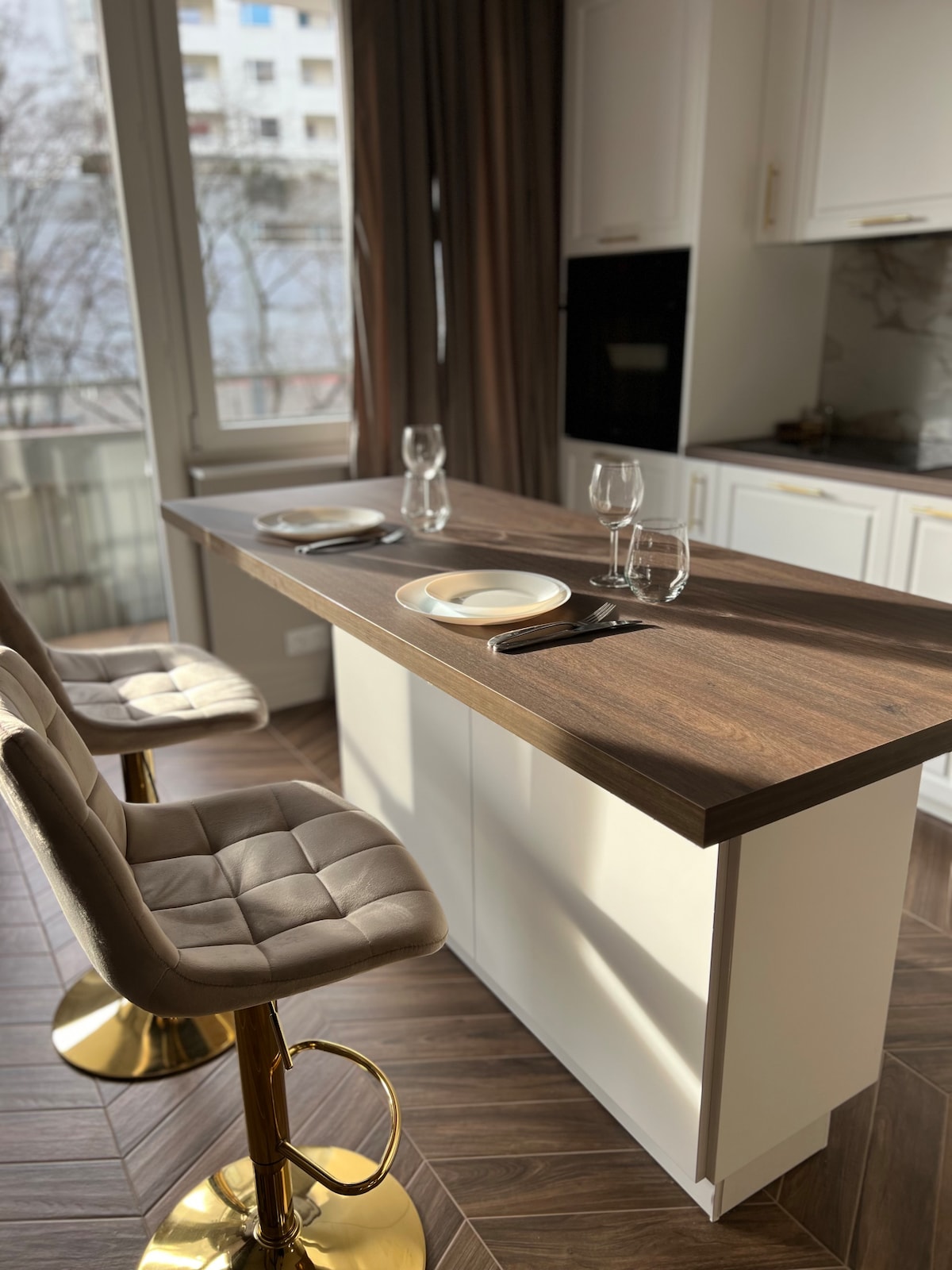 A modern kitchen island features a sleek wooden countertop with two place settings, including plates and glasses. Two stylish bar stools with gold bases are positioned at the island, complemented by large windows that allow natural light to brighten the space.