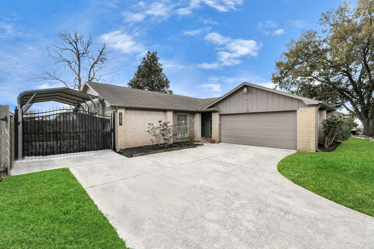 A single-story home is set on a spacious lawn, featuring a well-maintained driveway leading to a garage. A carport is visible, providing covered parking. The front yard includes small shrubs and a clear blue sky overhead, creating a welcoming entrance.