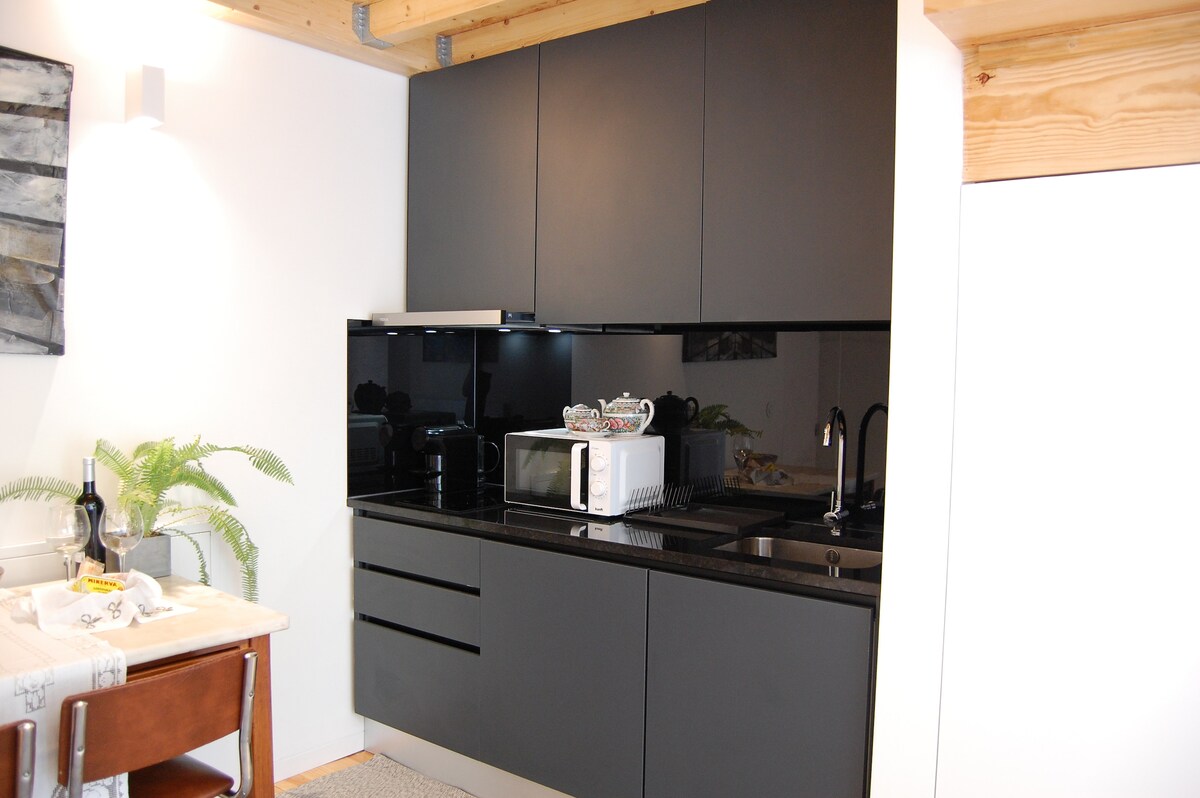 A modern kitchenette is presented with sleek black cabinets and a glossy backsplash. Essential appliances, including a microwave and sink, are neatly arranged. A small dining table is visible, accompanied by a chair and decorative elements, enhancing the functional design of the space.