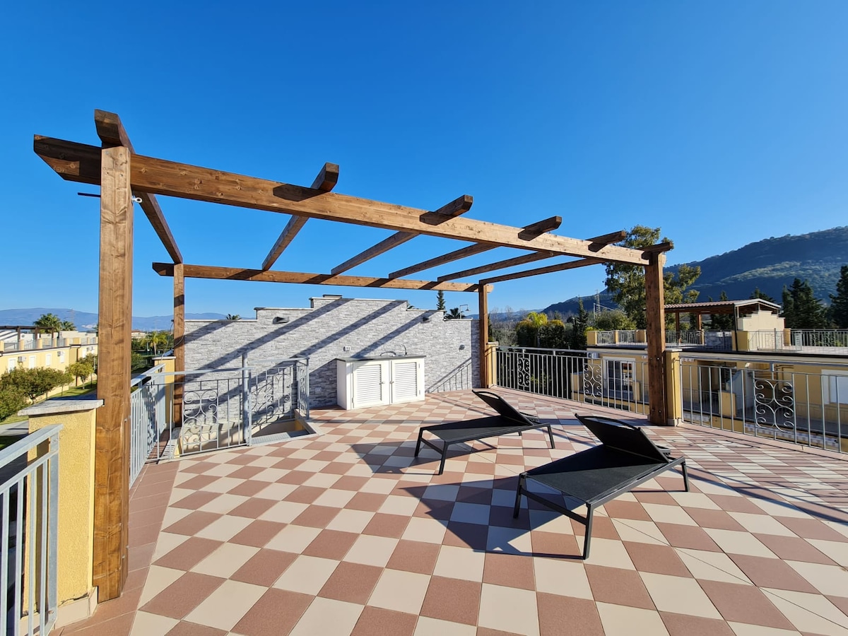 The rooftop solarium features a spacious tiled area with two lounge chairs positioned for relaxation. A wooden pergola provides shade, and distant mountains are visible in the background under a clear blue sky.