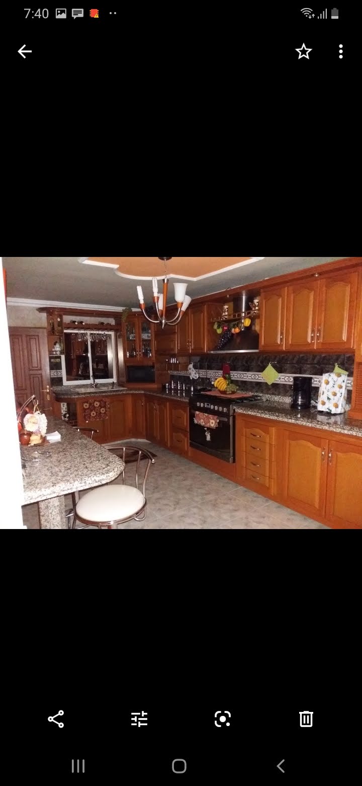 A modern kitchen features wooden cabinetry and sleek countertops. Stainless steel appliances are arranged along the walls, while a central island with seating is present. Decorative items and plants add a touch of color to the space, complemented by warm lighting from a stylish chandelier overhead.