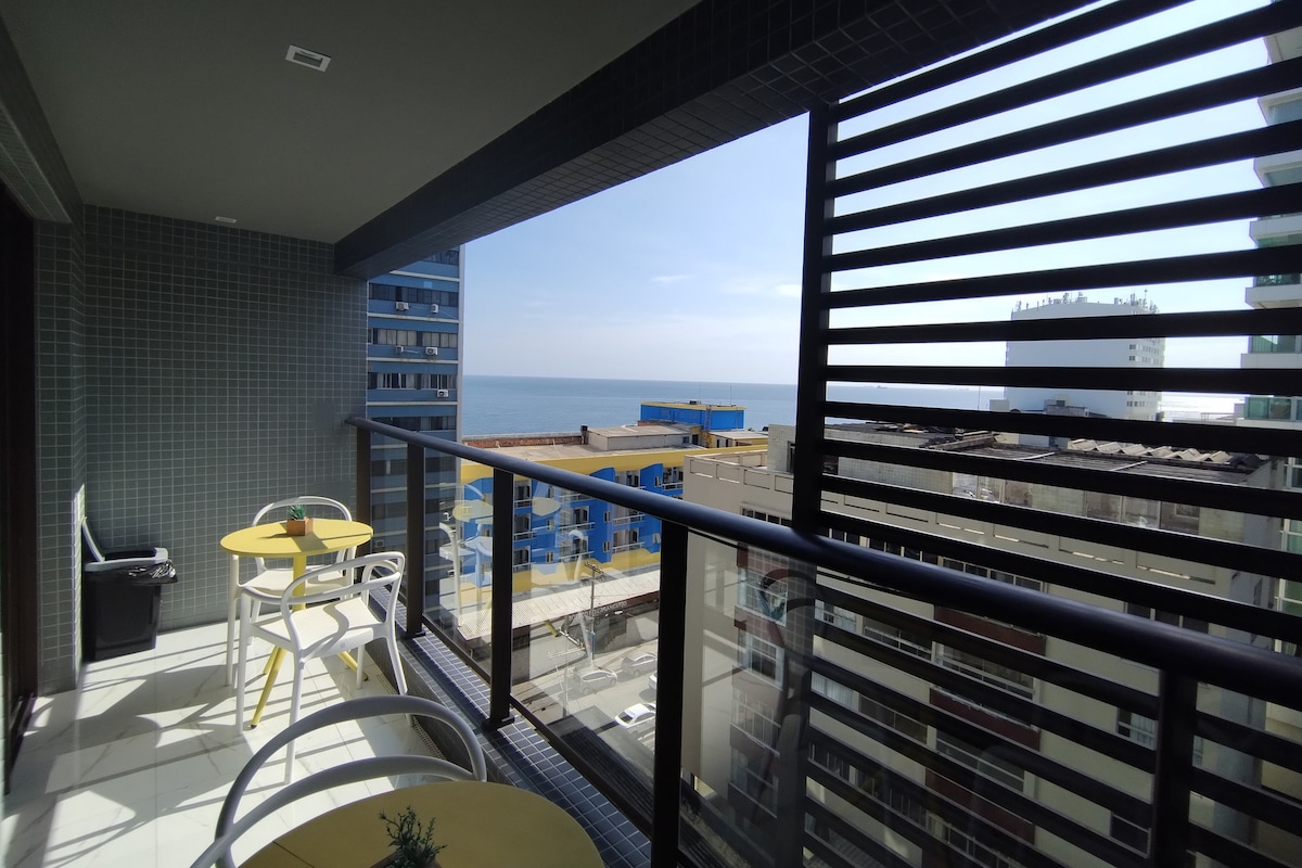 A balcony offers a view of the ocean, framed by sleek, modern railings. Two round tables with yellow tops and white chairs are positioned on the balcony, providing a bright and inviting space to relax. The horizon stretches above the water, with urban buildings visible in the foreground.