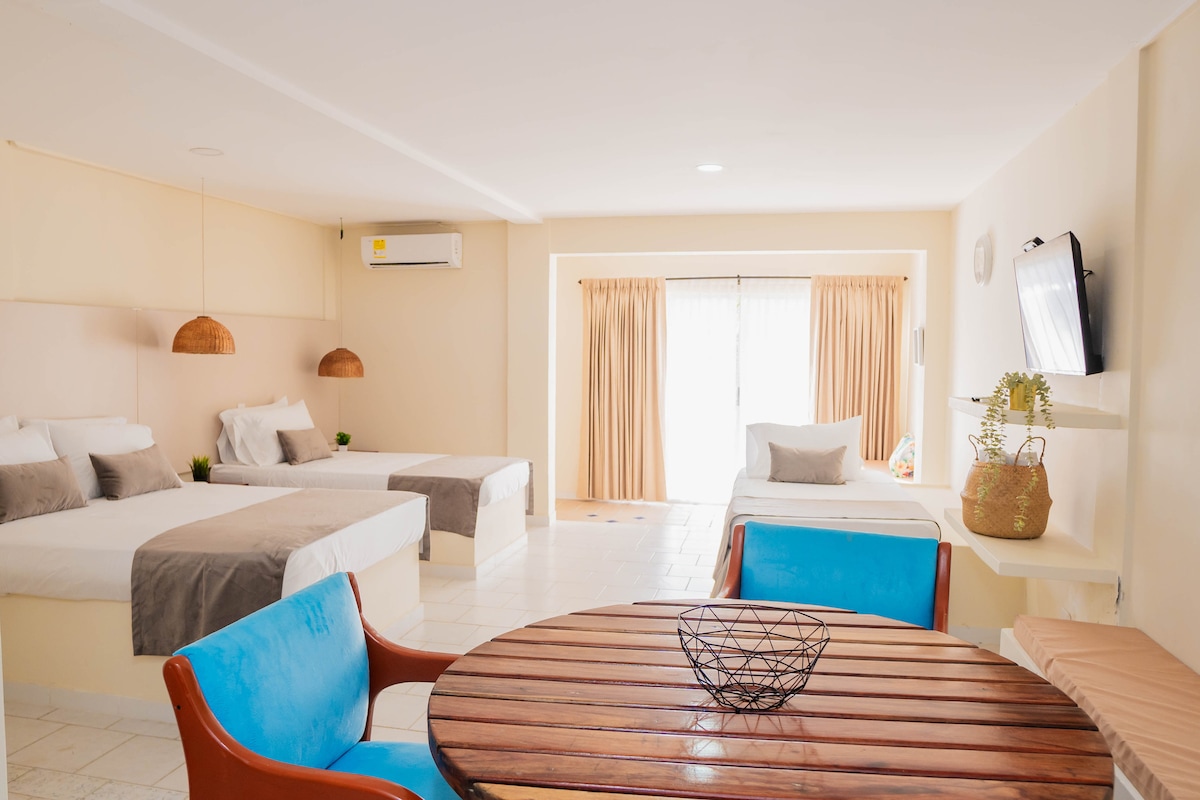 A spacious room is presented with two beds and a separate sleeping area featuring additional bedding. A round wooden table is accompanied by turquoise chairs. Large windows with light-colored curtains allow natural light to fill the space, creating an inviting environment.