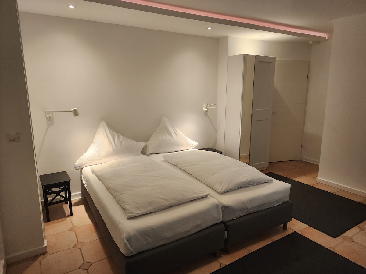 A modern sleeping area features a spacious bed with two large pillows positioned against a minimalist white wall. Adjacent to the bed, a small black bedside table is visible, and light fixtures provide soft lighting. The floor is covered with neutral-toned tiles.