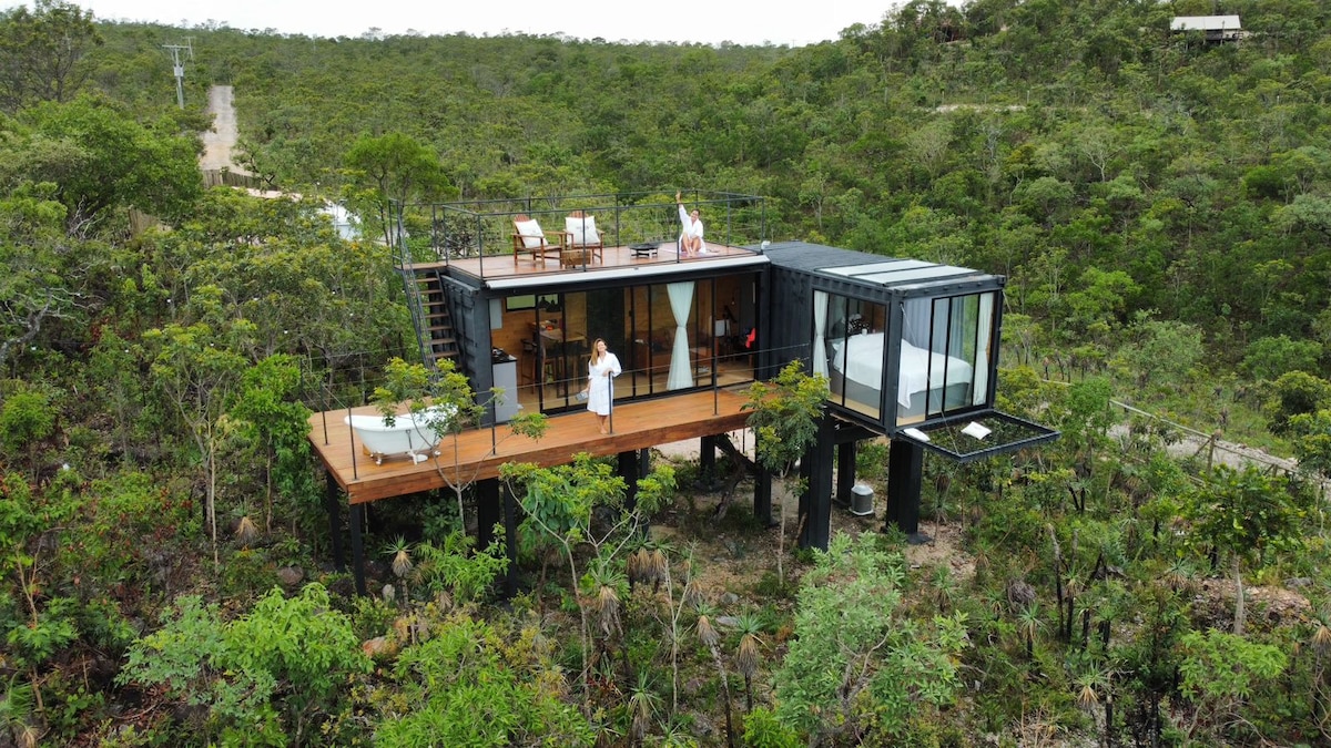 A modern structure elevated on stilts is surrounded by lush greenery, showcasing expansive glass walls that provide wide views of the natural landscape. A spacious deck features outdoor seating and a hot tub, while a bright sleeping area is visible through the glass.