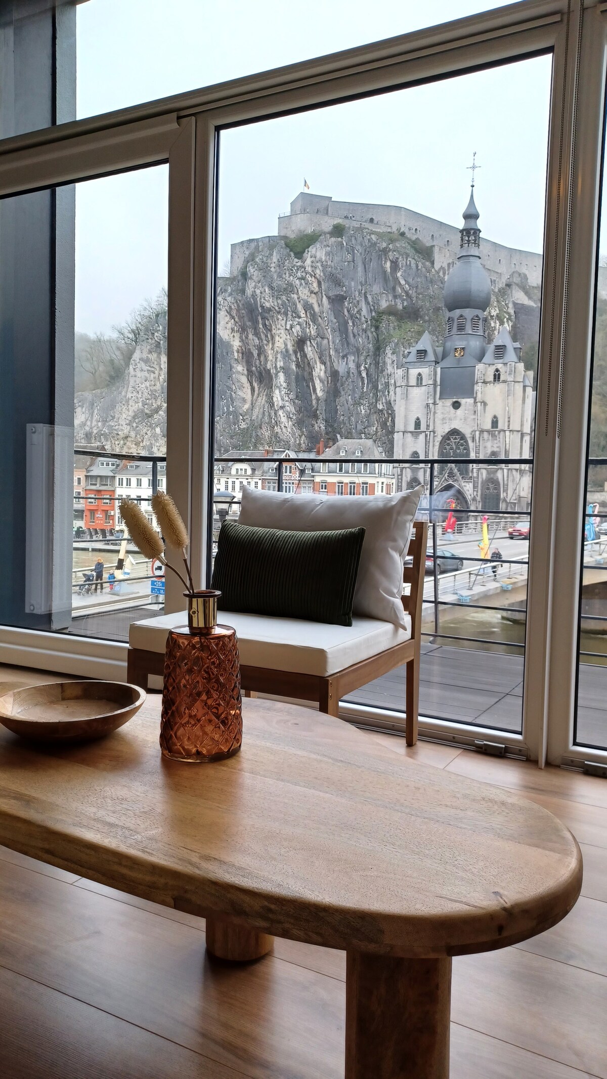 A cozy seating area features a wooden coffee table and a comfortable chair with a cushion. The expansive glass doors provide a panoramic view of the Citadel and Collegiate Church, which are framed against a backdrop of rocky cliffs and a vibrant town.