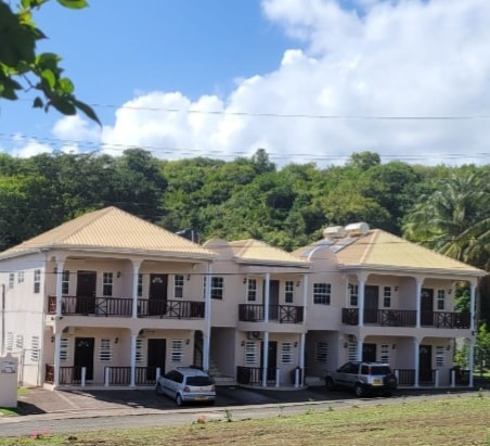 A two-story building with a light-colored exterior features multiple balconies. Lush greenery surrounds the property, with a clear blue sky overhead. Several parking spaces are visible, accommodating vehicles in front of the structure.