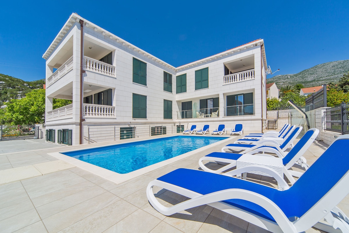 A modern apartment building is shown with a bright blue swimming pool in the foreground. Lounge chairs with blue cushions are arranged neatly around the pool area. The exterior of the building features light-colored stone walls and green window shutters, set against a clear blue sky.