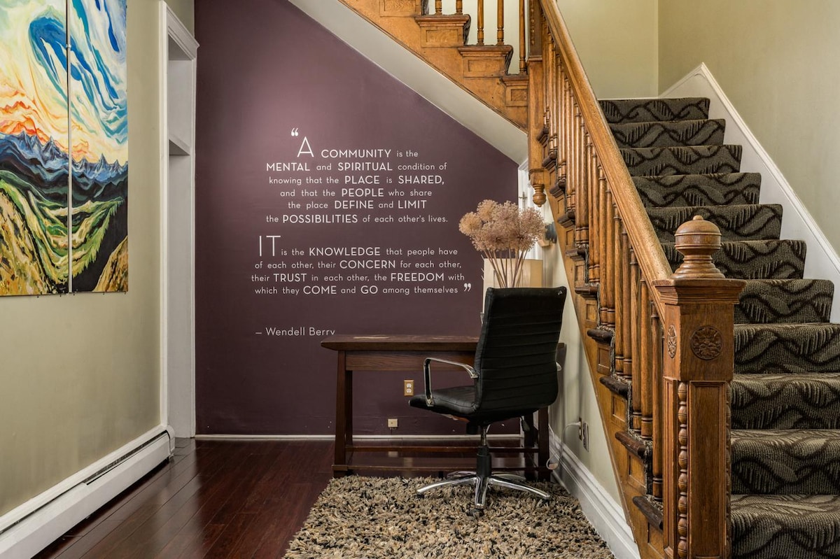 An inviting entryway features wooden stairs leading to the upper level. A plush area rug covers the floor near a stylish chair positioned against the wall. Inspirational quotes are displayed on the wall, enhancing the welcoming ambience of the space.