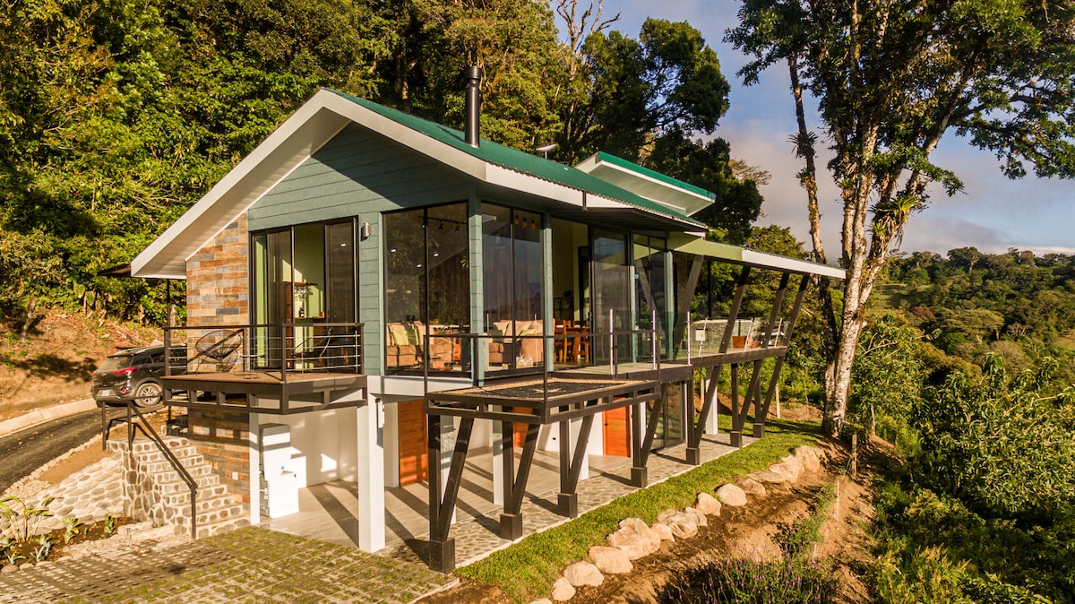 The modern house features large glass windows and a spacious veranda, offering panoramic views of the surrounding tropical landscape. The structure is accented by a stone façade and a green roof, set against lush greenery and a clear sky.