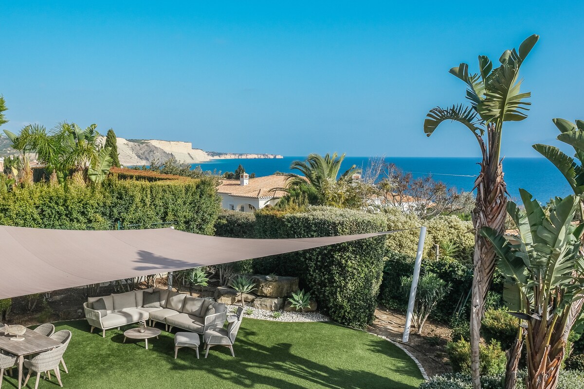 A well-maintained garden showcases a spacious area with a shaded seating arrangement under a canopy. Lush greenery surrounds the space, providing a sense of privacy. The view opens up to a stunning coastline, with the ocean glistening in the distance.