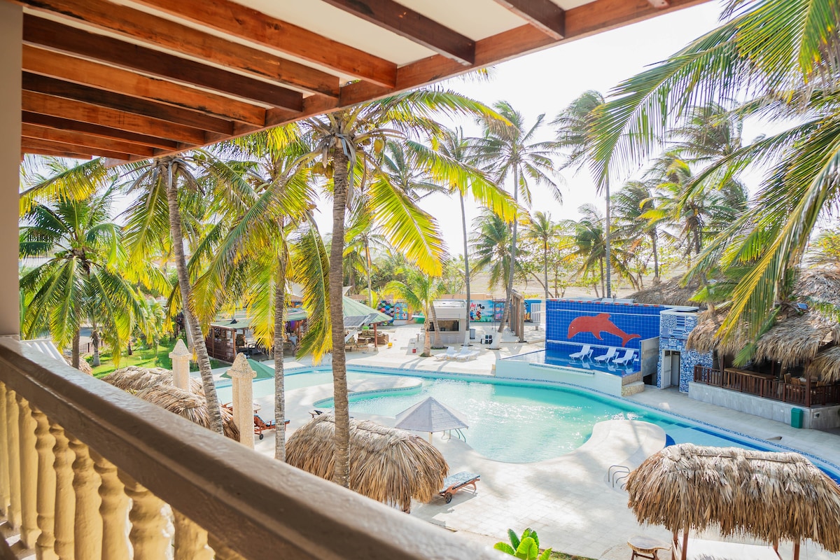 A view from a balcony showcases a vibrant pool area surrounded by tall palm trees. Lounge chairs are positioned near the water, and thatched umbrellas offer shade. The pool is set against a backdrop of tropical landscaping, enhancing the serene environment.