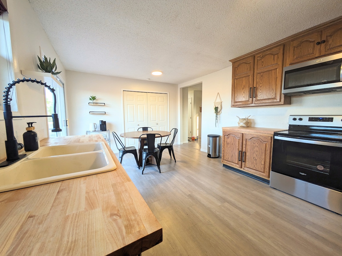 A well-equipped kitchen features wooden cabinetry and stainless steel appliances. A dining table with four black chairs is situated near a window, allowing natural light to brighten the space. The countertops are made of light wood, creating an inviting working area.