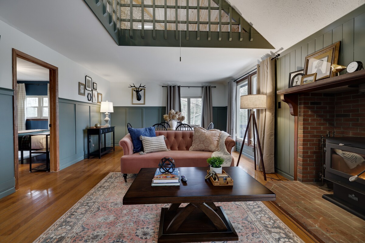 A welcoming living area features a pink sofa paired with a wooden coffee table, set on a patterned area rug. A fireplace is situated against a brick wall, while large windows allow natural light to fill the space. The room includes decorative wall art and a floor lamp.