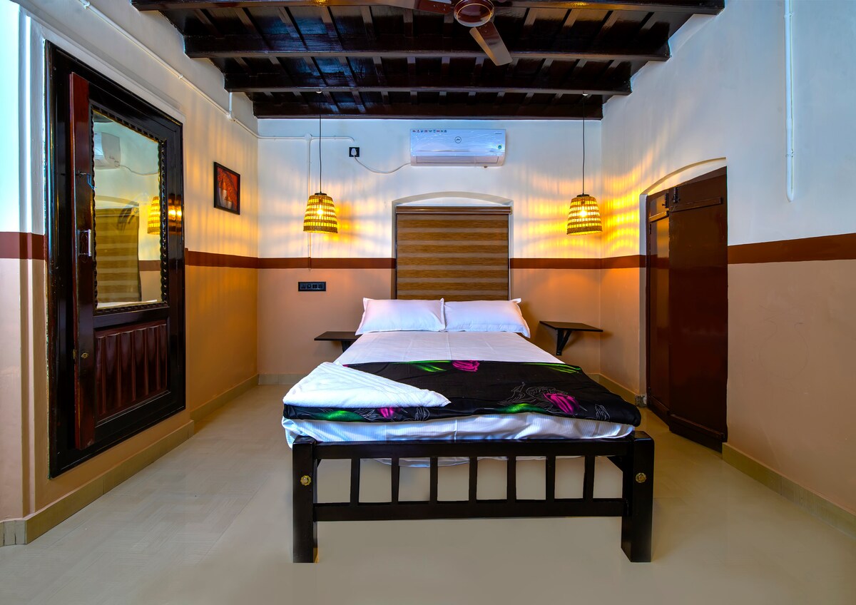 The room features a spacious bed with a decorative blanket, set against a backdrop of textured walls and wooden ceiling beams. Soft lighting is provided by two pendant lights, while a window and door ensure natural light fills the area. A small table is positioned next to the bed.