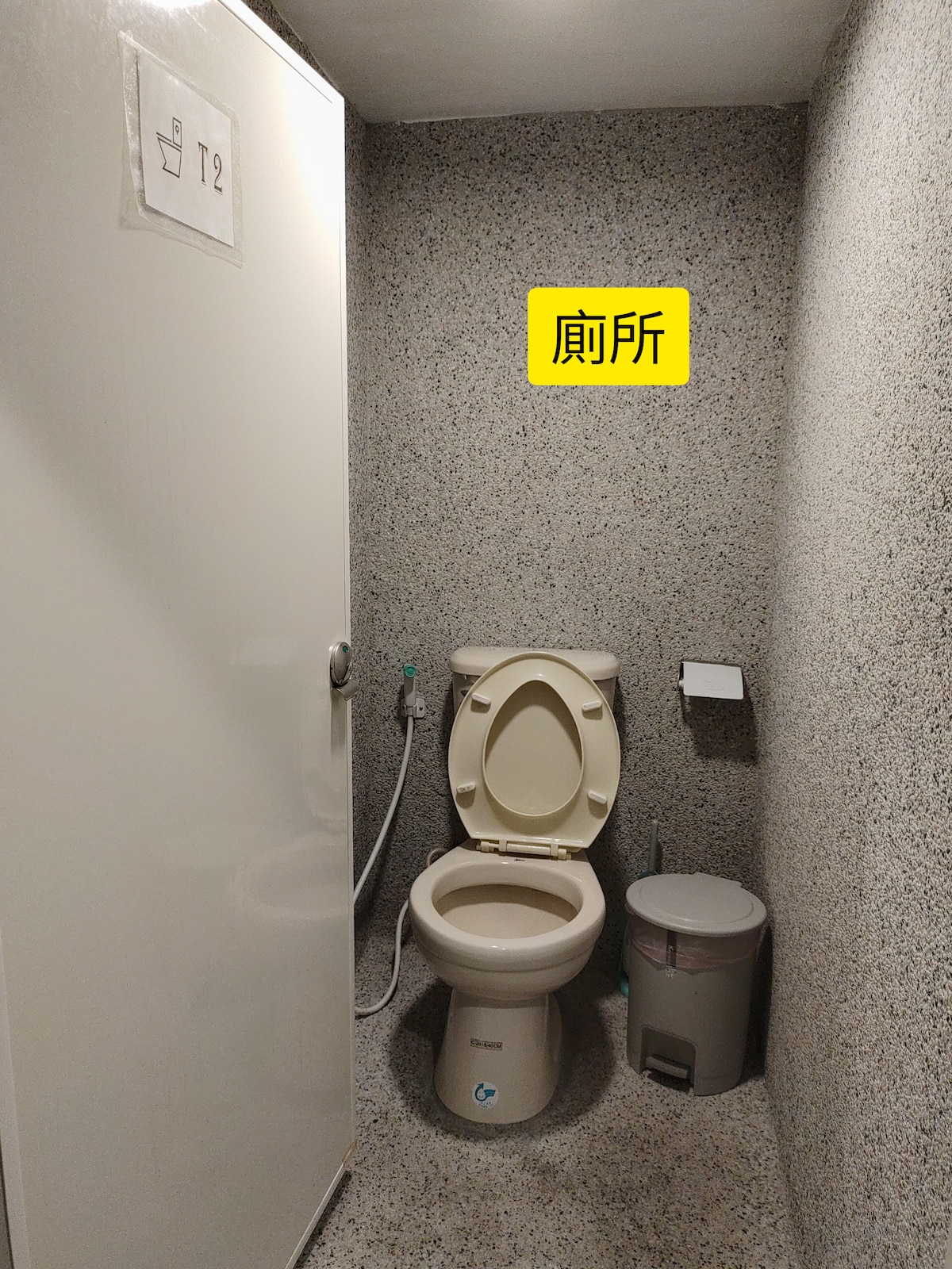 A functional bathroom space features a white toilet and a small waste bin. Textured gray walls create a clean backdrop. A door with a simple design provides privacy.
