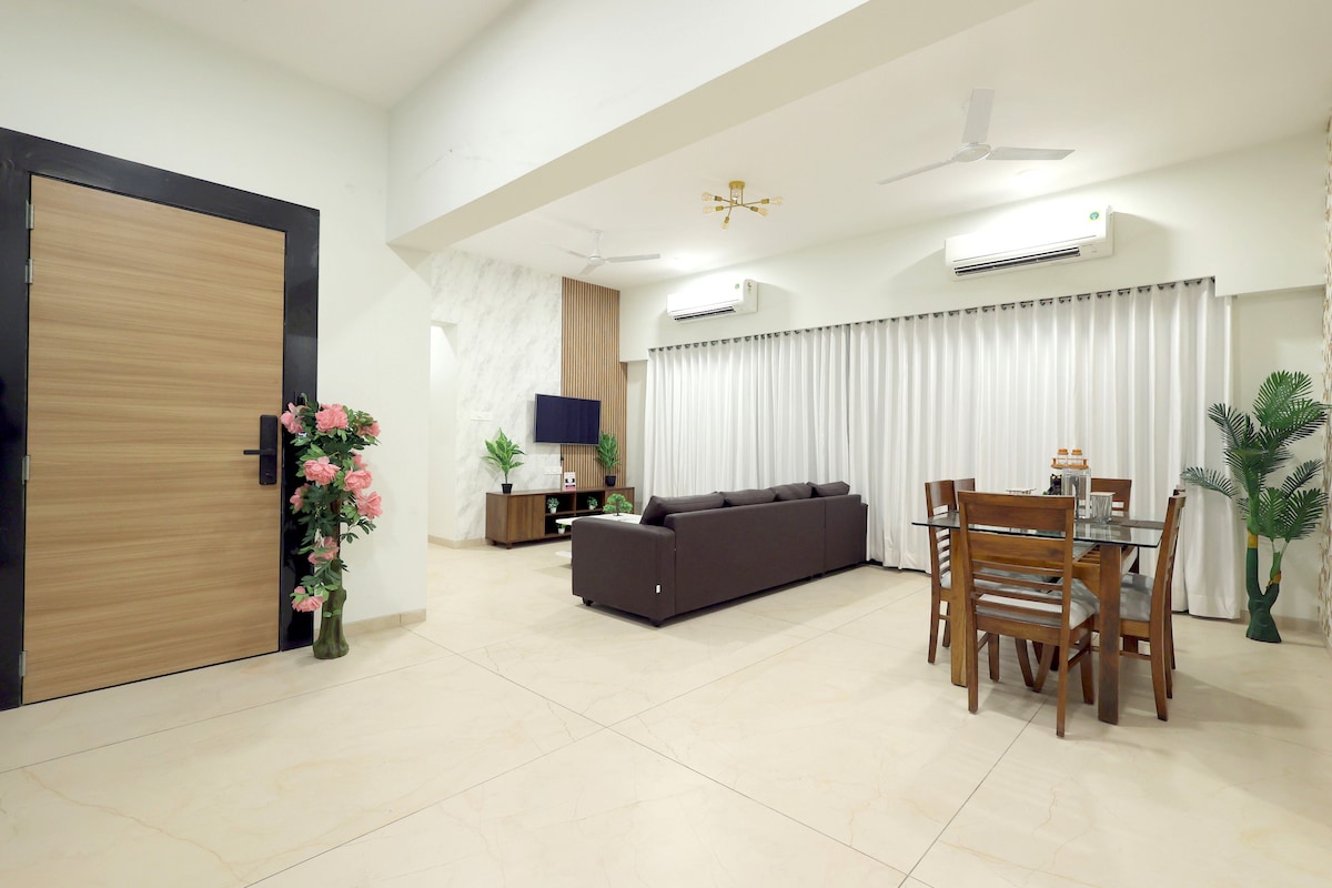The spacious living area features a modern design with a comfortable sofa, wooden dining table, and a flat-screen television. Large windows with light-colored curtains allow natural light to fill the room, while potted plants add a touch of greenery.