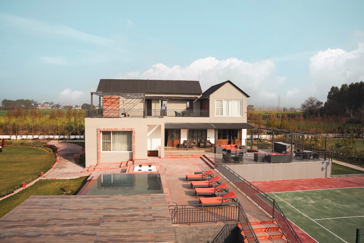 A two-story farmhouse features modern architecture with large glass windows. The outdoor area includes a private pool and a well-maintained tennis court, surrounded by green lawns. Lounge chairs are arranged by the pool, creating a spacious and inviting atmosphere for relaxation.