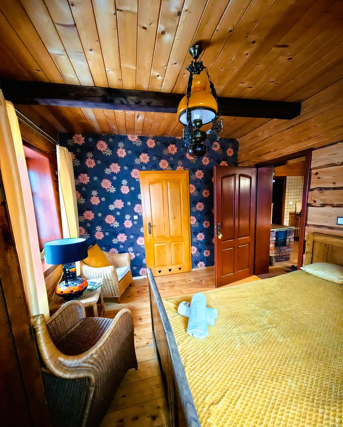 A cozy bedroom features warm wooden walls and a vintage lamp, casting soft light. Floral wallpaper adds a touch of color, while a spacious bed with a golden cover is complemented by a wicker chair and a small side table near a window with light curtains.