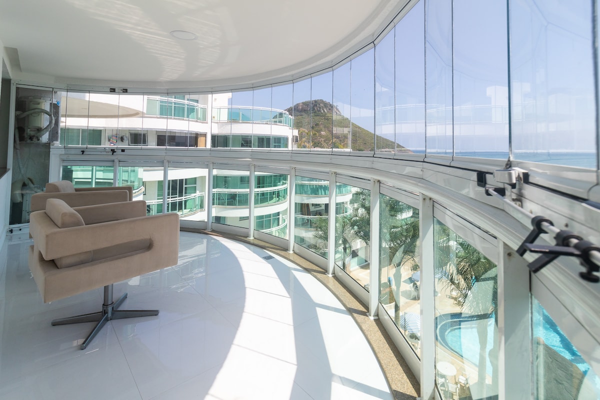 A spacious balcony offers panoramic views of the beach and surrounding landscape. Fixed glass panels provide a clear sightline without obstruction. A light-colored chair is placed near the window, creating a relaxing spot to enjoy the scenery.