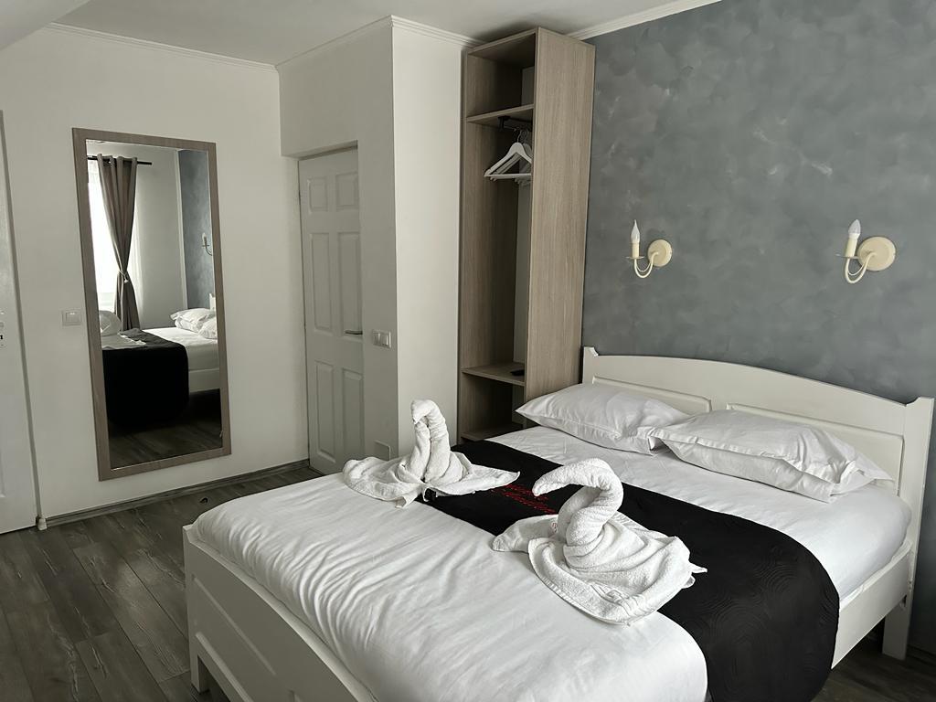 The sleeping area features a spacious bed with white linens and a black decorative runner. Two towel swans adorn the bed. A mirror and a small clothing rack are positioned against the wall, while soft curtains frame the window, allowing natural light to brighten the space.