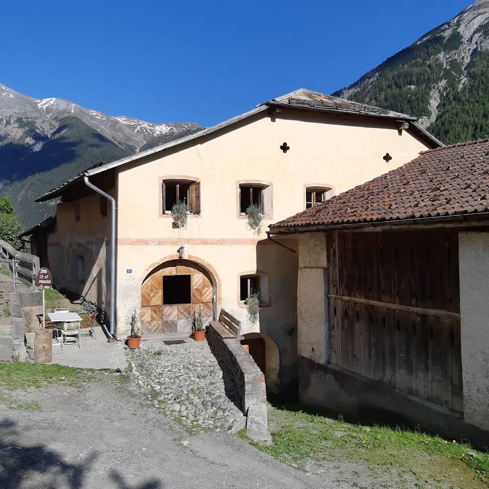An old Engadine house is depicted, showcasing a rustic exterior with light-colored walls and wooden features. A stone pathway leads to the entrance, flanked by greenery. The surrounding mountains create a serene backdrop, highlighting the house's connection to nature.