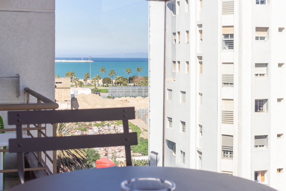 A view from the balcony reveals a scenic coastline, featuring the sea and palm trees in the foreground. The space includes a small table and chair, providing a spot to enjoy the sea breeze and the surrounding urban landscape.