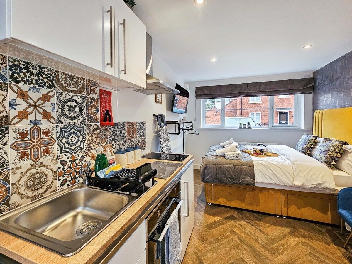 A compact studio apartment is displayed, featuring a fully equipped kitchen with patterned tiles and modern appliances. A sleeping area with a double bed is visible in the background, accented by colorful cushions and a wall-mounted television. Natural light enters through a window, enhancing the space.