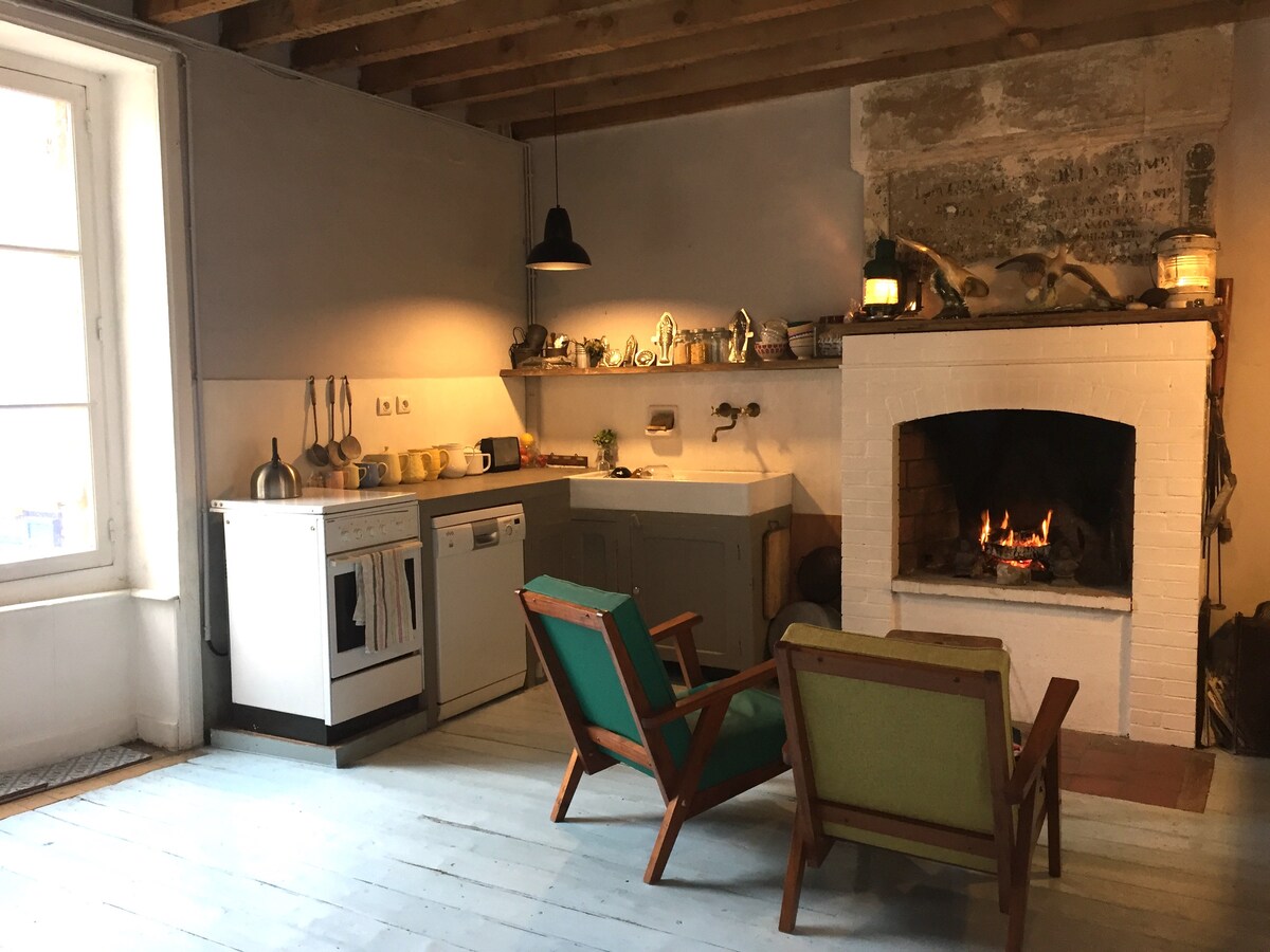 A kitchen area features white cabinetry and modern appliances, including a stovetop and sink. A cozy fireplace is set against an exposed brick wall, with a fire gently burning inside. Two colorful mid-century chairs add seating options, complemented by wooden beams overhead.