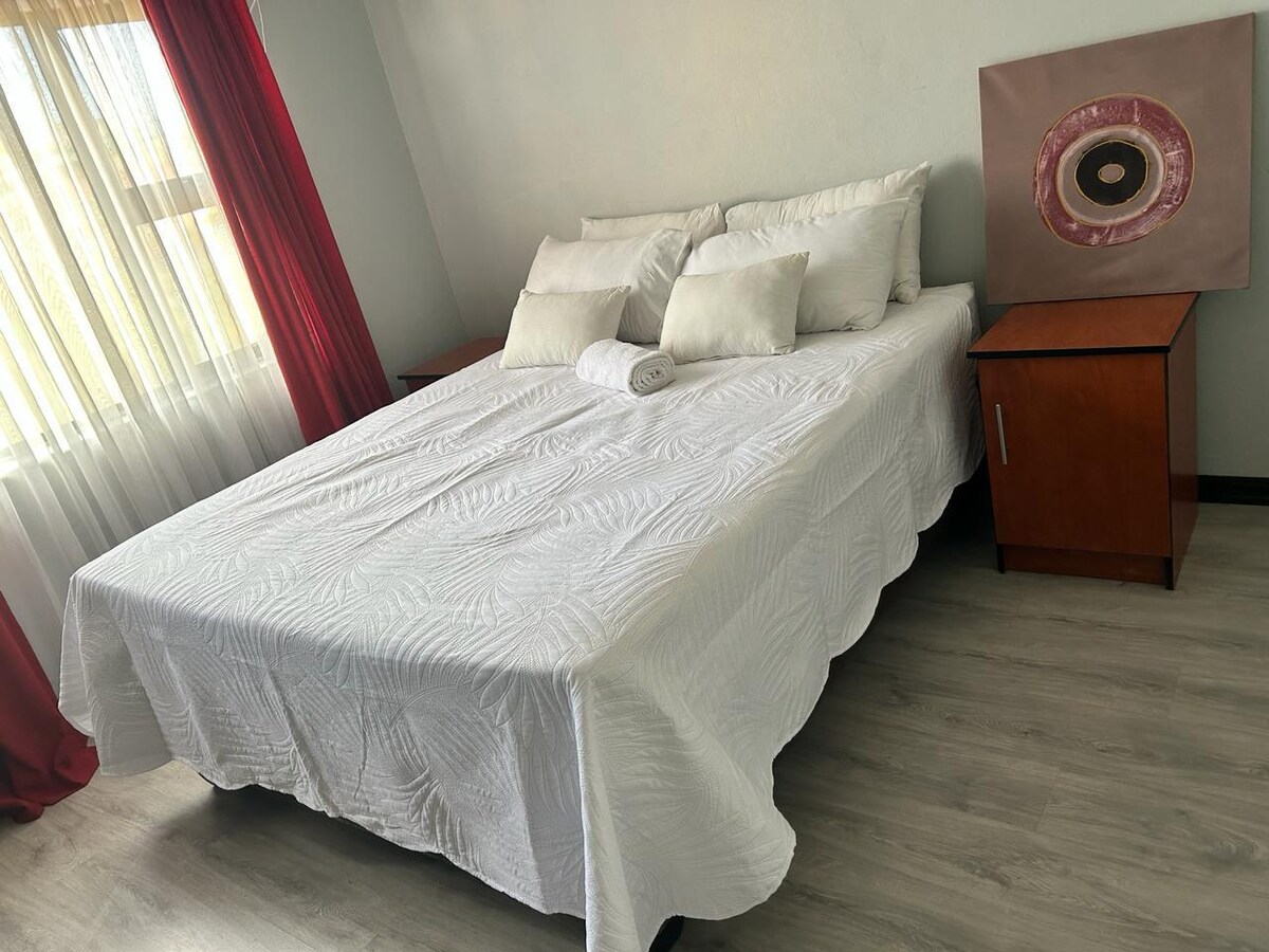 A spacious bed is topped with white linens and several decorative pillows. Beside the bed, a wooden nightstand provides a contrast to the light bedding. Natural light filters through sheer curtains, brightening the room with a soft glow.