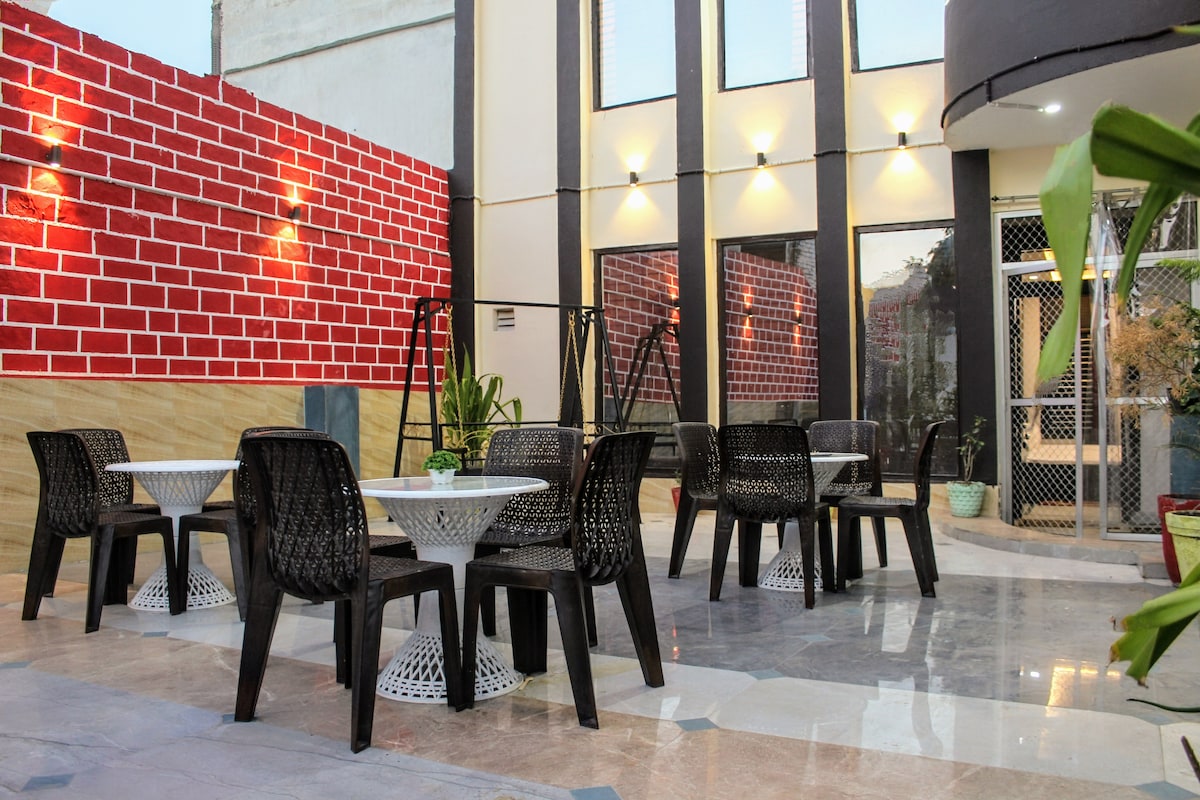An outdoor seating area is presented with multiple black chairs surrounding round tables. Brightly lit walls create a vibrant backdrop, while a swing adds an inviting touch. The smooth flooring reflects the surrounding greenery, contributing to a relaxing ambiance.