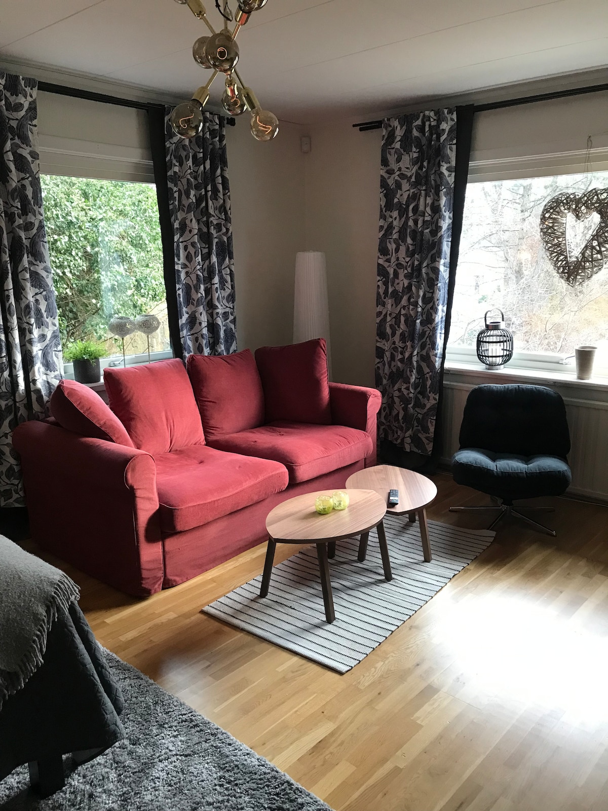 The living area features a red couch positioned near large windows dressed with patterned curtains. A simple round table sits on a striped rug, complemented by an accent chair. Natural light illuminates the space, revealing wooden flooring and decorative plants.