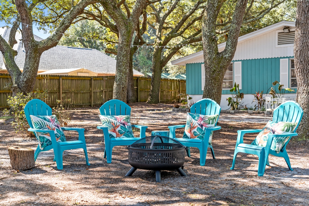 A cozy outdoor seating area is arranged around a fire pit, featuring four vibrant blue chairs with tropical-patterned cushions. Tall oak trees provide shade over the gravel ground, offering a relaxed setting for gatherings. A quaint bungalow is visible in the background.