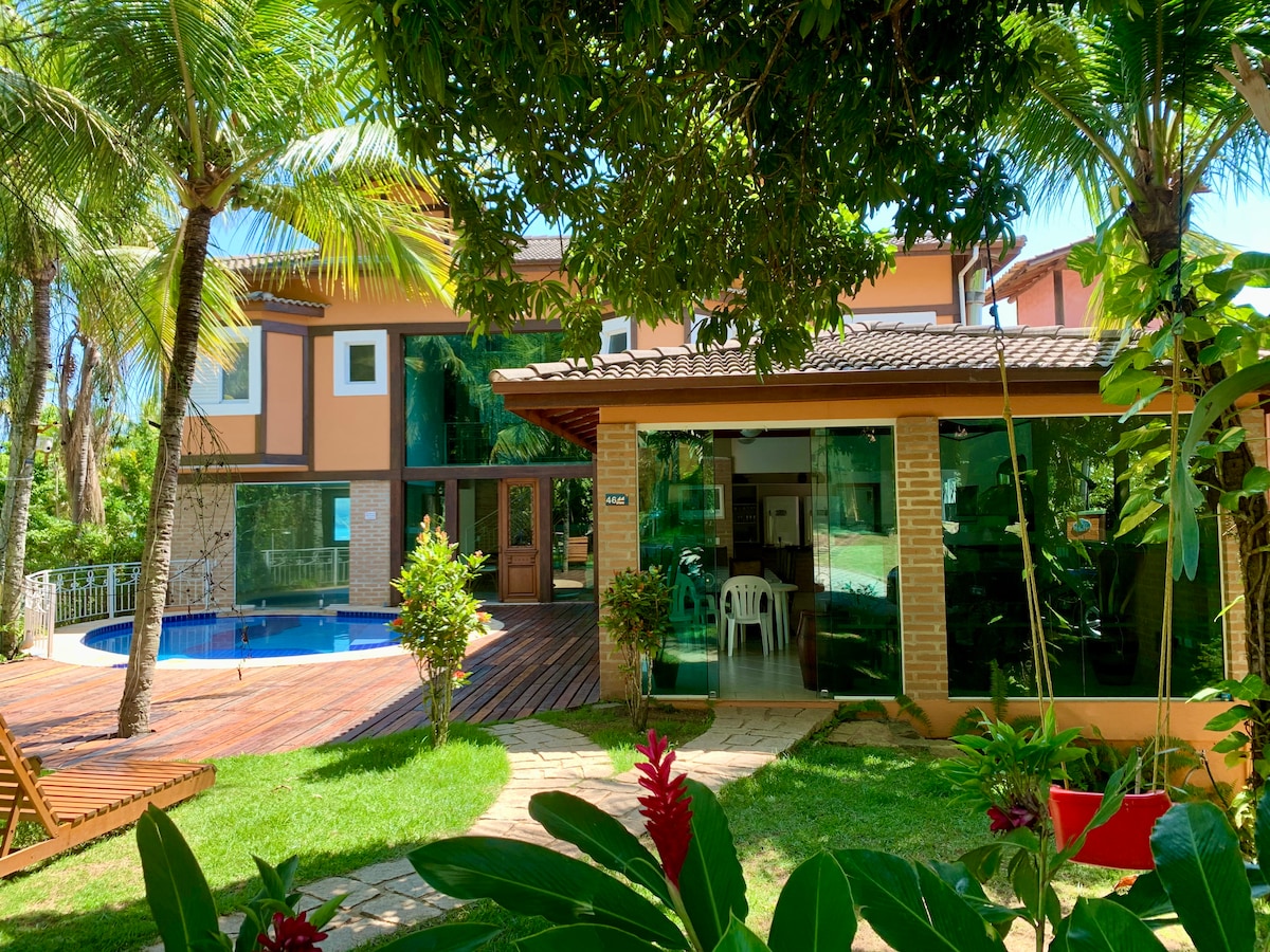 The exterior of the house features a sunlit pool surrounded by tropical greenery. Large glass windows offer views into the indoor spaces, while a covered patio with white chairs provides a comfortable area for relaxation. Lush plants and trees enhance the natural ambiance.