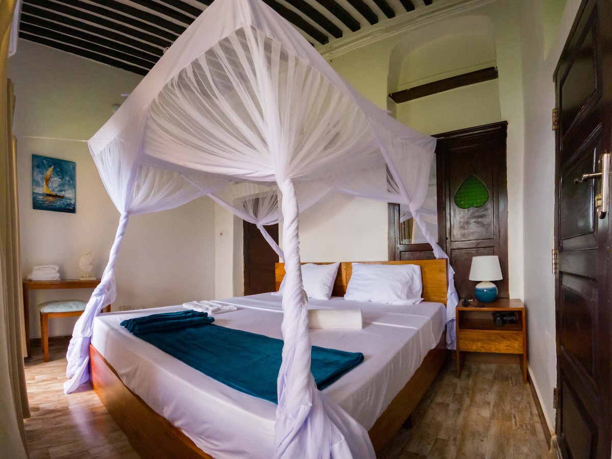 A spacious bedroom features a canopy bed draped in sheer white fabric. Soft, cool-toned linens are accented by a teal blanket at the foot. Natural light fills the room, highlighting wooden furnishings, including a bedside table and a small desk. Traditional elements are subtly integrated throughout.