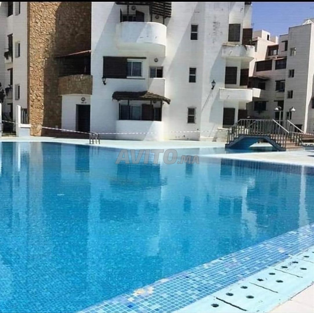 An outdoor swimming pool is prominently featured, reflecting the clear blue sky. Surrounding the pool, white buildings are visible, with balconies overlooking the area. Sun loungers may be arranged nearby, creating an inviting space for relaxation.
