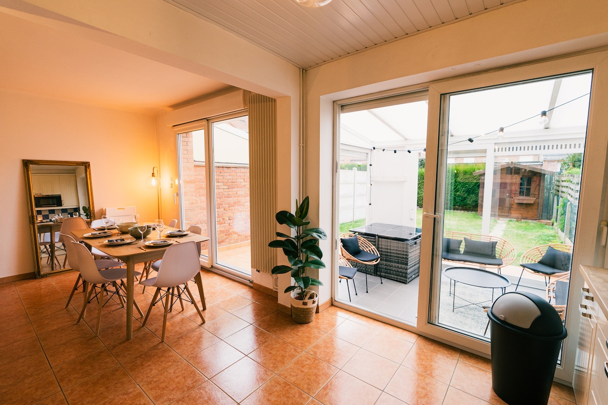 A dining area features a wooden table set for six, surrounded by white chairs. Large glass doors lead to a private garden, which is visible through the open space. Natural light fills the room, complemented by a potted plant near the window.